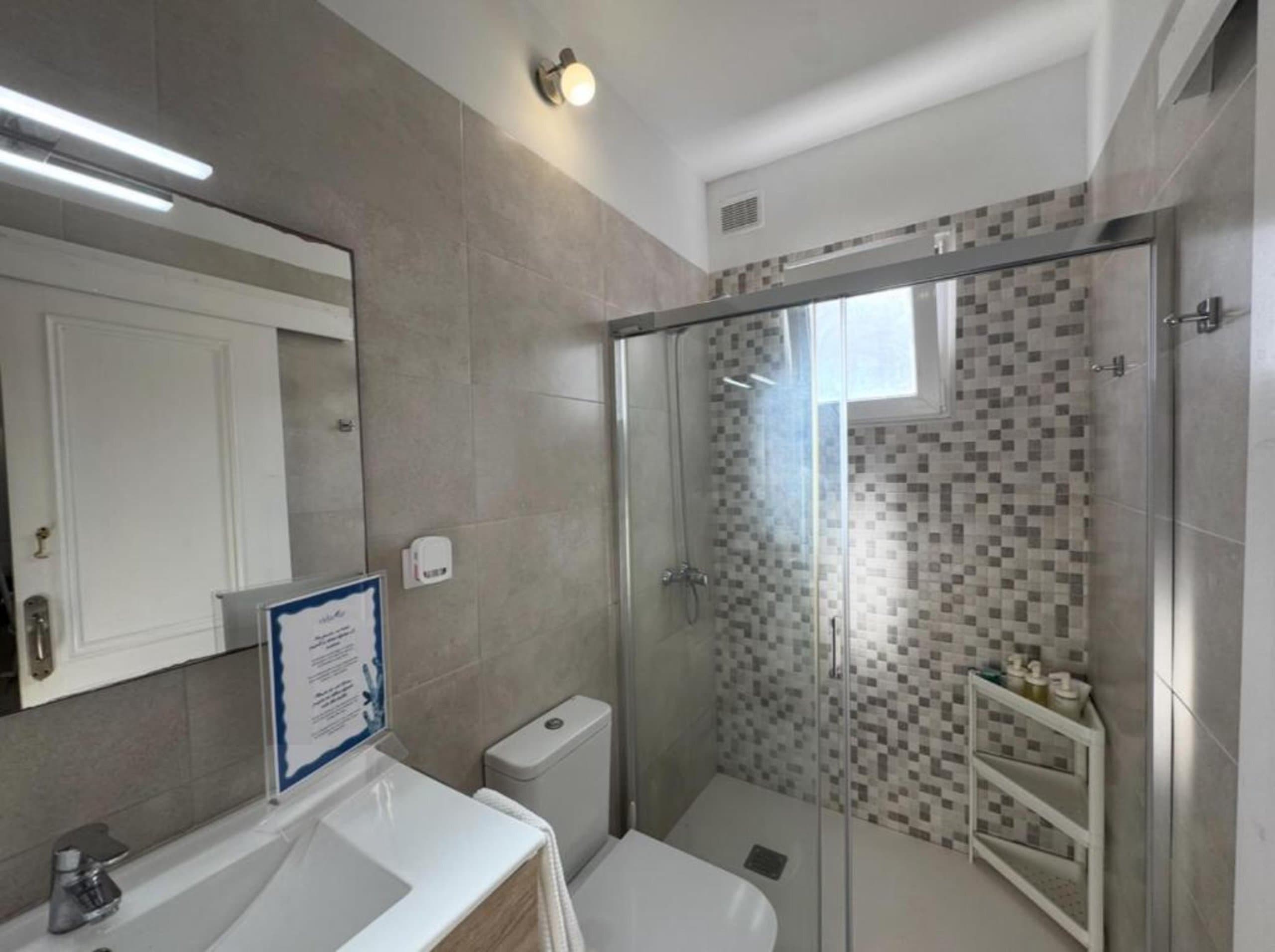Baño moderno y luminoso con ducha de mampara, azulejos decorativos, espejo amplio, lavamanos blanco y acabados contemporáneos. Diseño limpio y funcional.